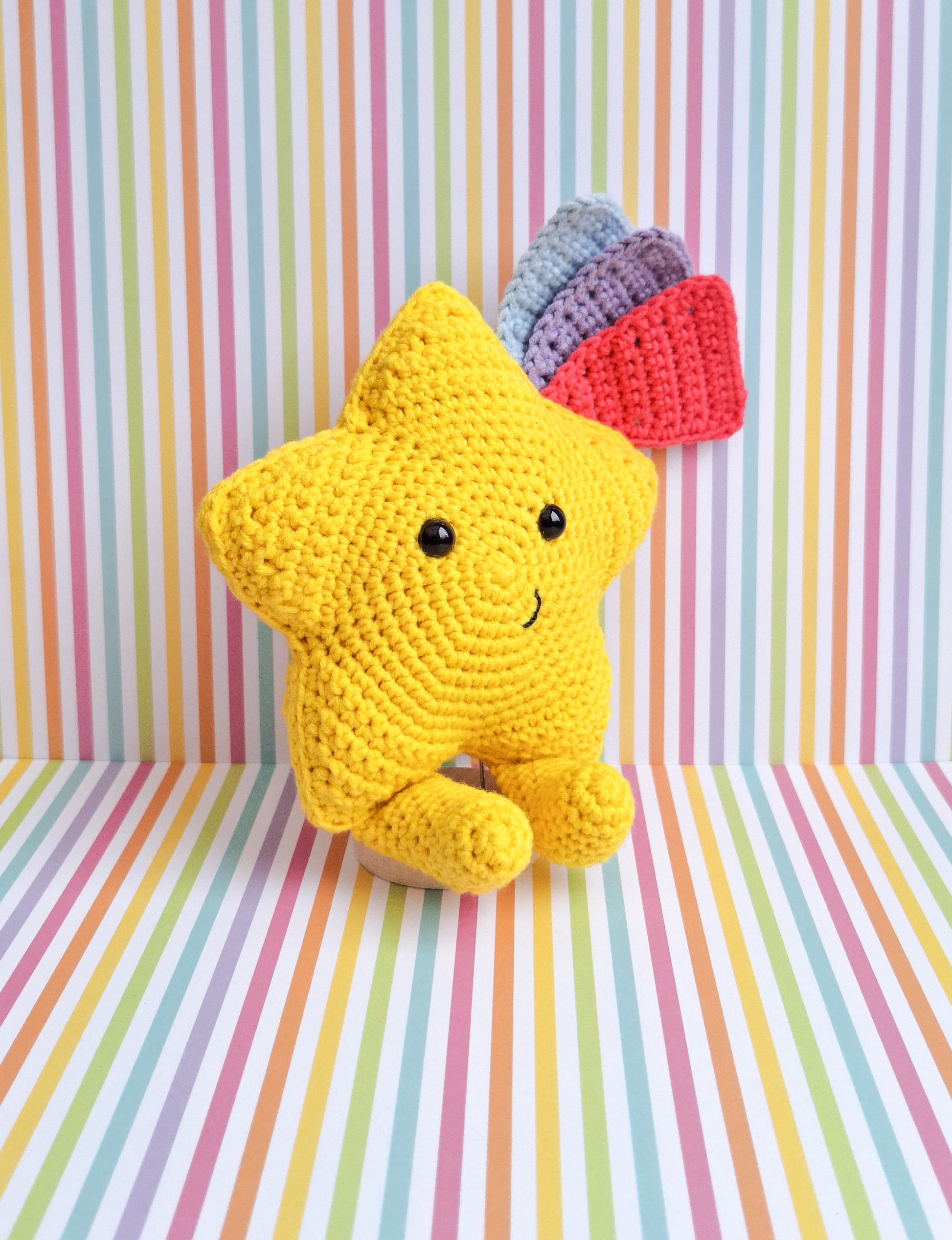 A kawaii star made from the advertised crochet pattern. A crochet star made in yellow cotton yarn with feet and 3 shooting star tails in blue, pink and purple