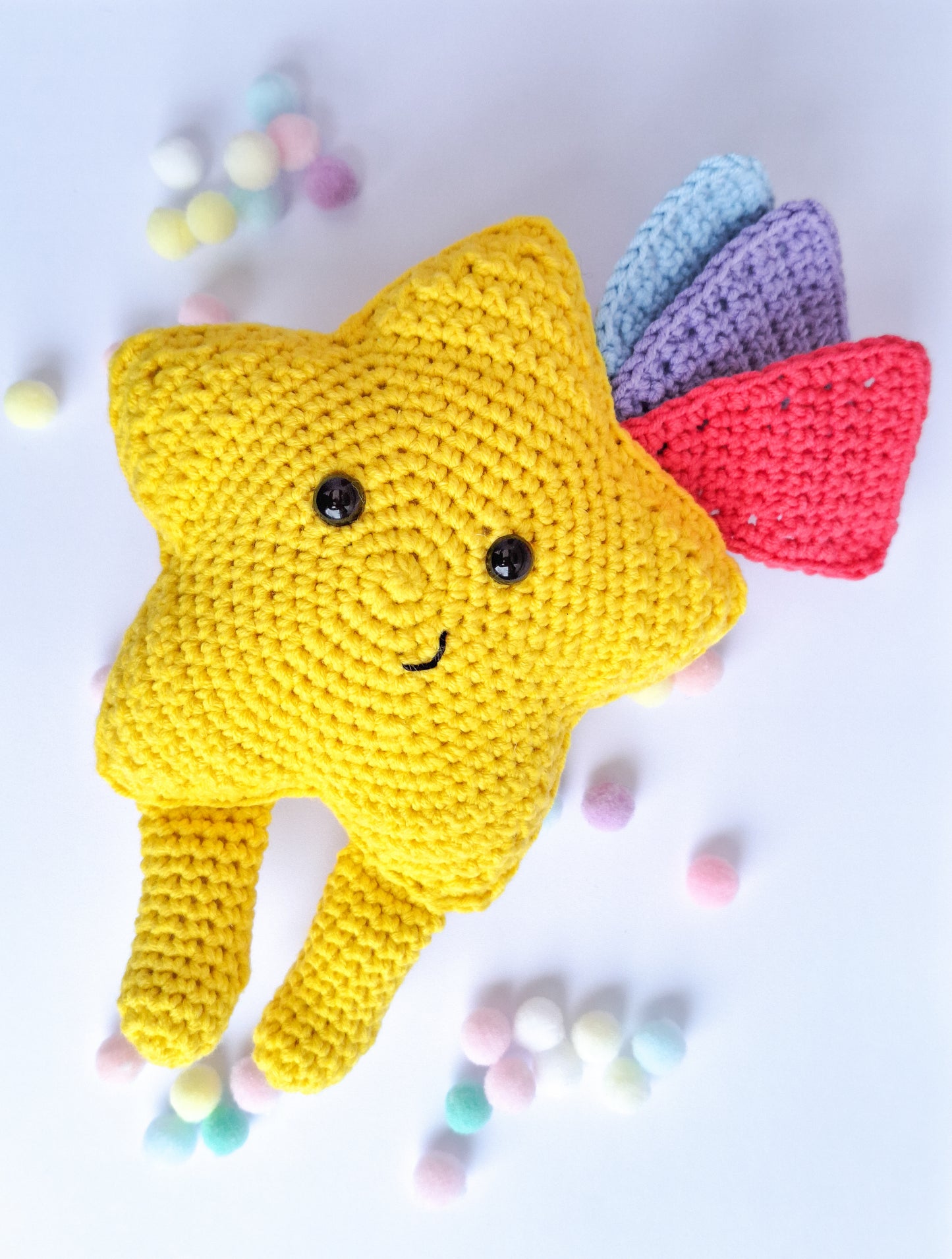 A kawaii star made from the advertised crochet pattern. A crochet star made in yellow cotton yarn with feet and 3 shooting star tails in blue, pink and purple